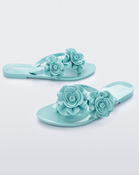 An angled front and side view of a pair of green Mini Melissa Harmonic Garden flip flops with flowers on the straps.