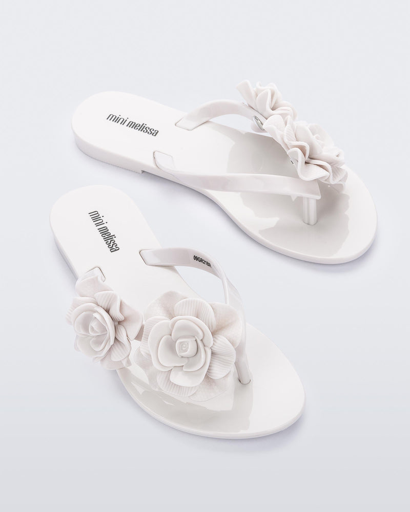 An angled top view of a pair of white Mini Melissa Harmomic Squared Garden flip flops with flowers on the straps.