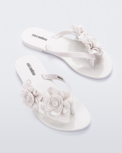 An angled top view of a pair of white Mini Melissa Harmomic Squared Garden flip flops with flowers on the straps.