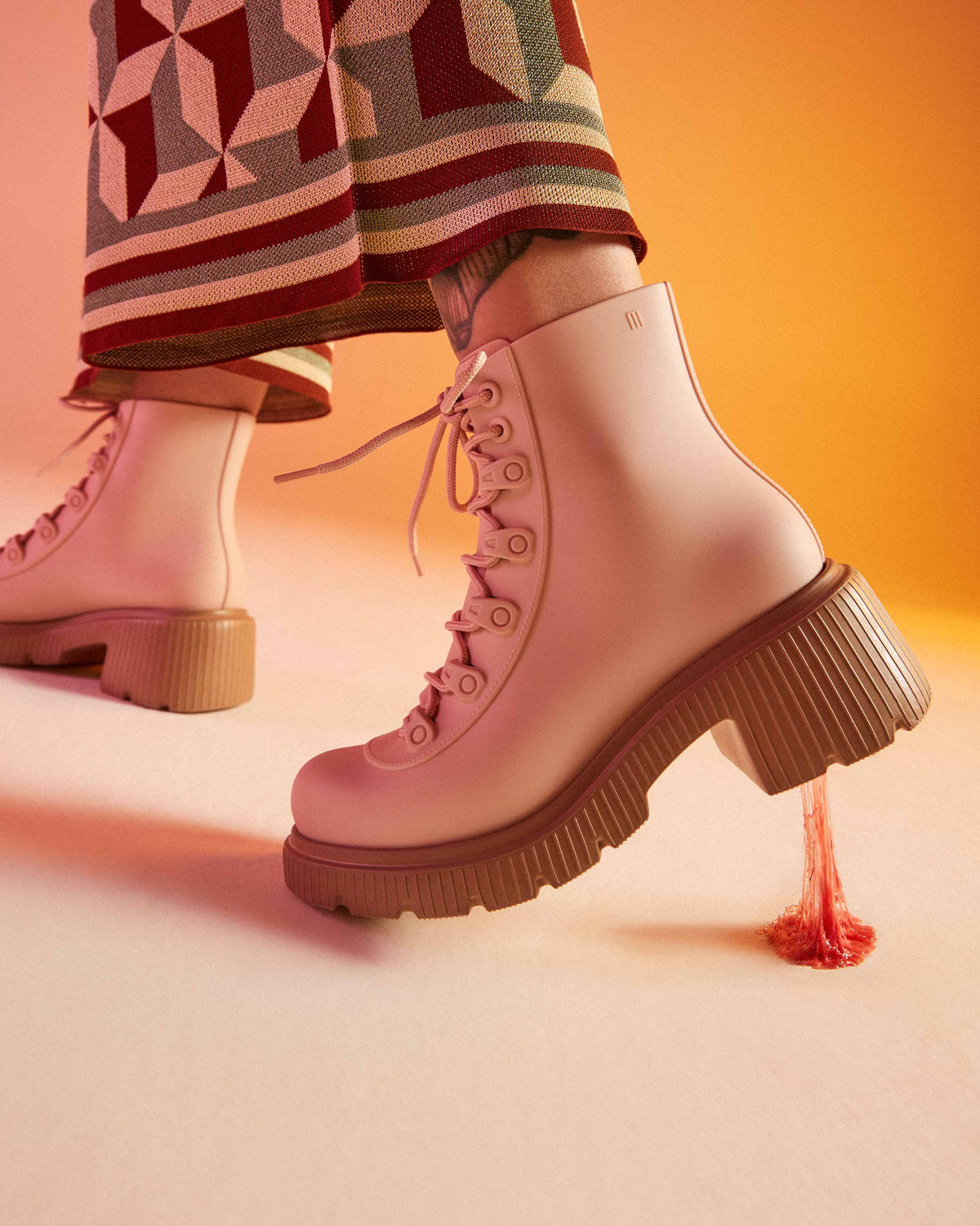 A model's legs wearing patterned pants and a pair of brown/pink Melissa Cosmo boots with pink base, laces and a brown heel sole, with a piece of gum stuck on the bottom.