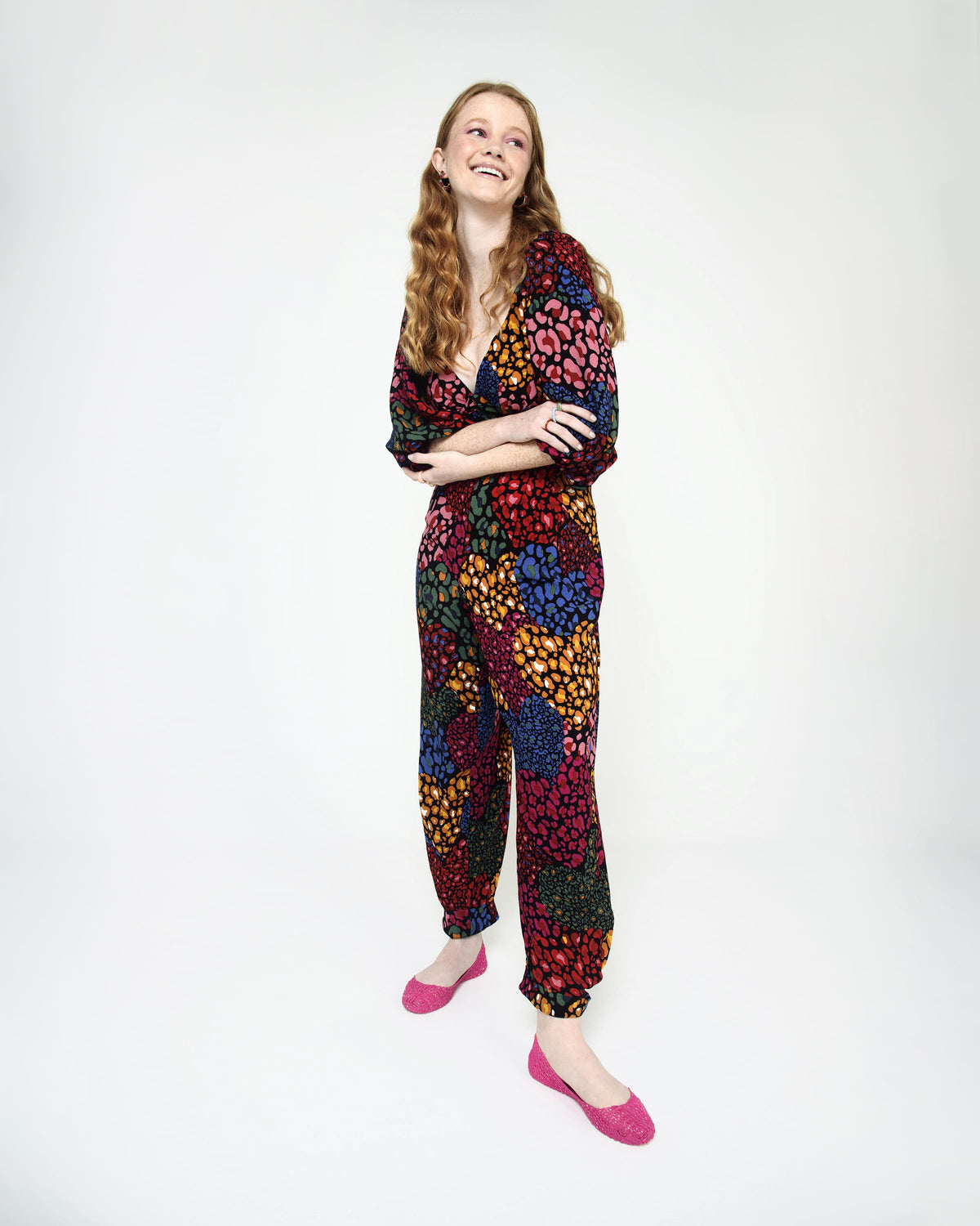 A woman standing with her arms crossed wearing a multicolored pattern jumpsuit with a pair of bright pink Melissa Campana flats.