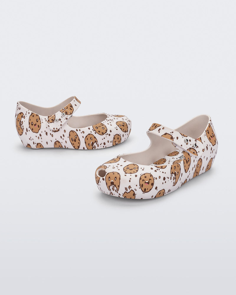 Angled view of a pair of Mini Melissa Ultragirl flats in candy print. The print is an all over white / beige chocolate chip cookie print. The shoe has a single velcro strap across the front of the foot.