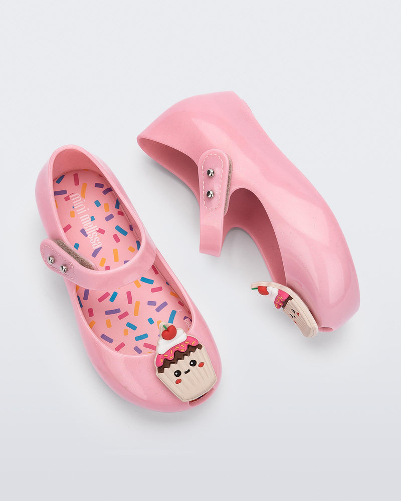 Top and side view of a pair of Mini Melissa Ultra Girl Candy ballet flats in pink glitter colorway. The shoe has a single strap on the top of the foot with a double snap closure. The shoe has a 3D printed cupcake applicque with smiling face on the top of the toe. The colors of the applique are red, brown and white. The insole of the shoe has a multicolored sprinkle print.