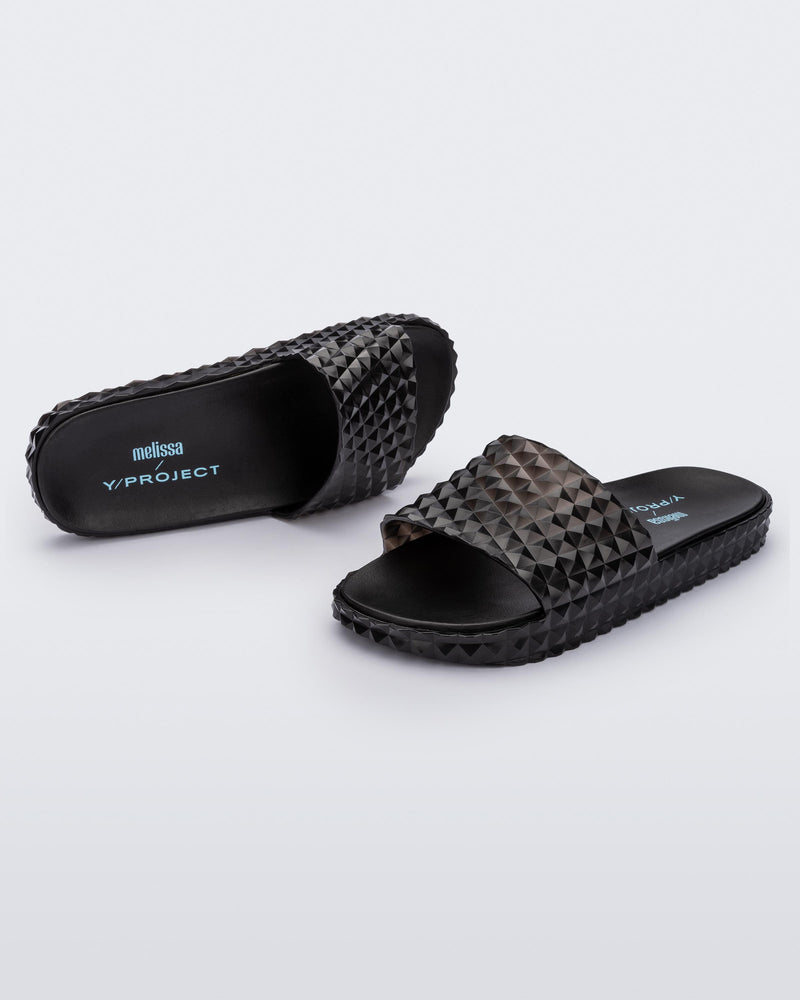 An angled front and top view of a pair of transparent black Melissa Court Slides with a checkered pattern texture.