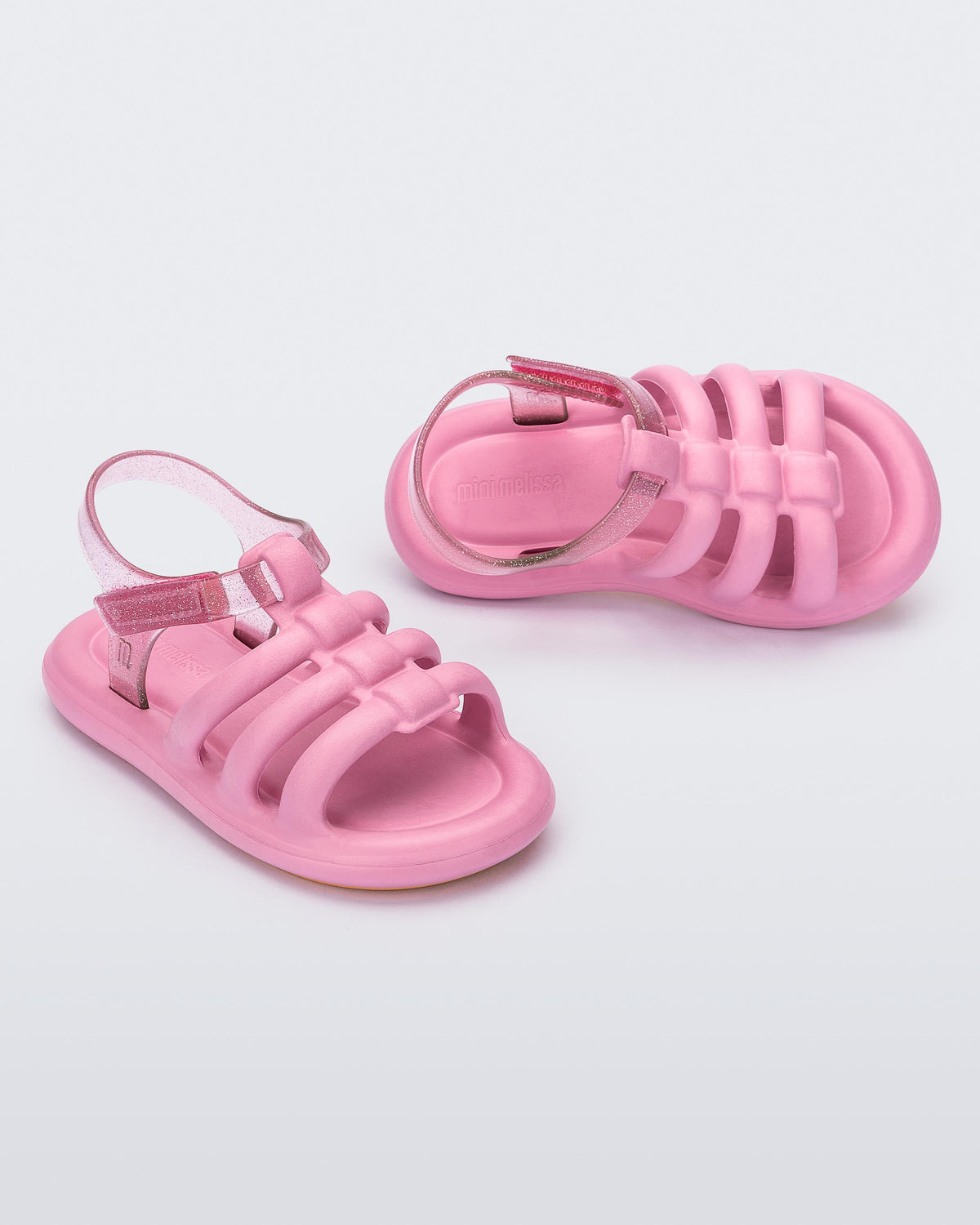 An angled front and side view of a pair of pink Mini Melissa Freesherman sandals with pink and clear glitter straps.