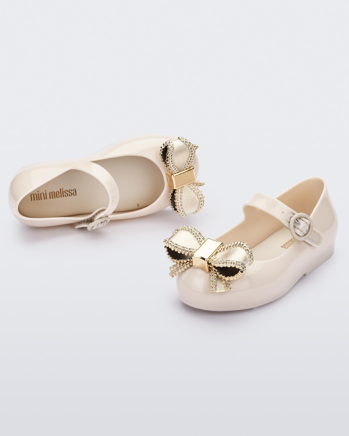 Top and angled view of a pair of beige Mini Melissa Sweet Love baby mary jane flats with a metallic gold bow.