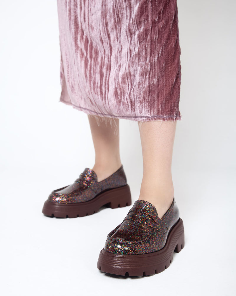 Closeup of a model wearing a pair of Melissa Royal loafers in glitter multicolor with a brown platform sole.