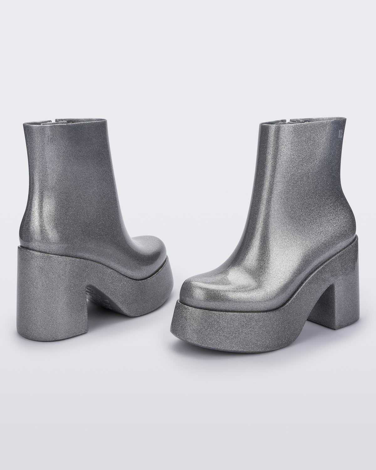 Angled and back view of a pair of silver glitter Melissa Nubia platform heel boots.