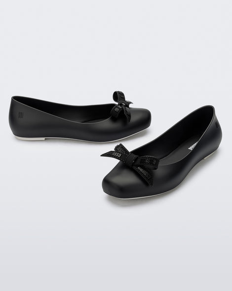 Angled view of a pair of Melissa Aura flats in black with bow details on a light gray background
