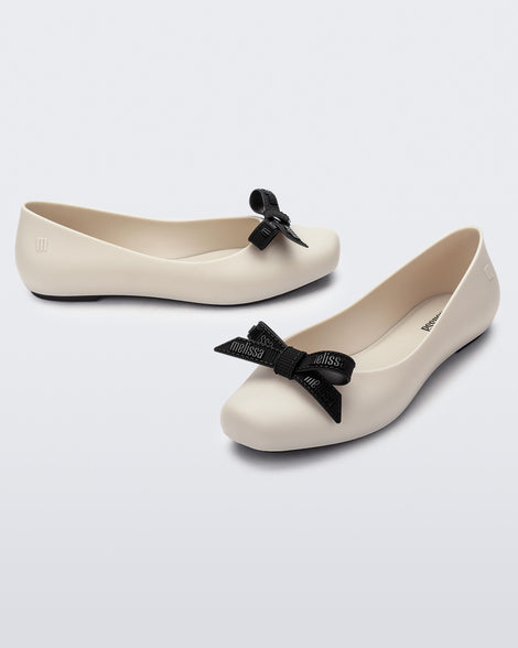Angled view of a pair of Melissa Aura flats in beige with black bows on a light gray background