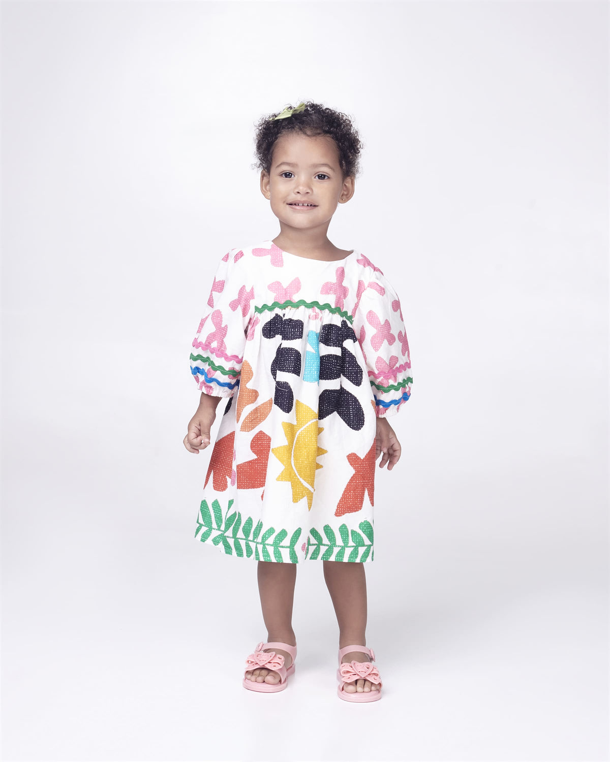 A baby model posing for a picture in a white patterned dress and a pair of glitter pink Mini Melissa Mar Sandal Heart sandals with a glitter pink heart bow detail on the front strap and an ankle strap