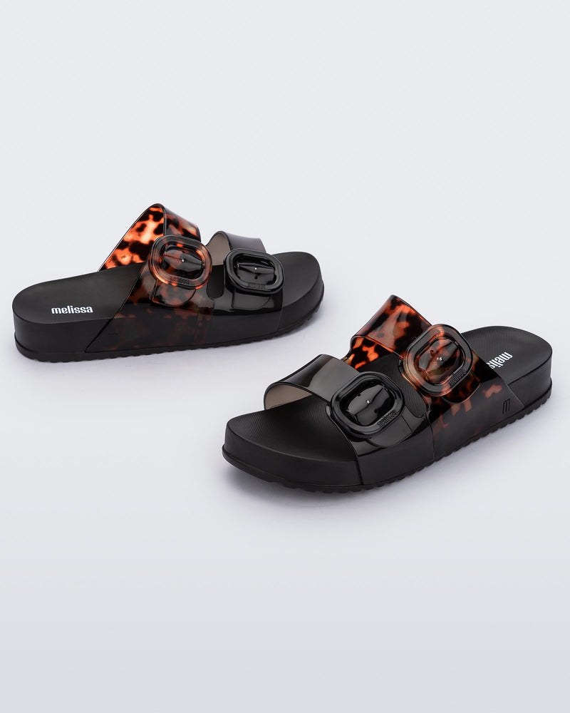 Side view of a pair of Black/Clear Tortoise Melissa Cozy slides with two black and transparent tortoise straps with a buckle detail.