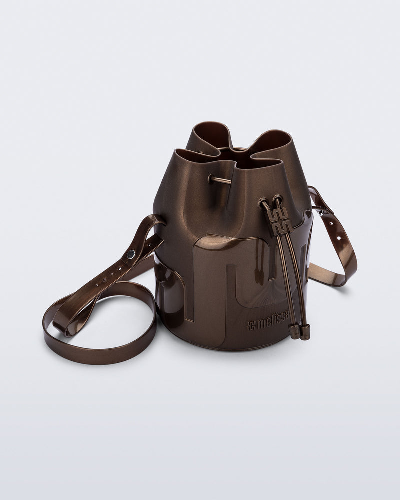Angled view of a Melissa Station bucket bag in Bronze. The bag has an adjustable shoulder strap and a drawstring top closure.