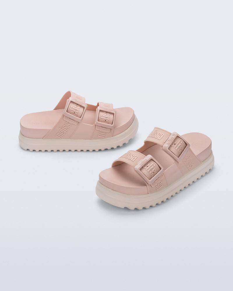 Angled view of a pair of melissa cozy platform slide sandals. There are two buckle straps that go across the foot with the M Lover (melissa logo) print embossed into the straps. The buckles are located on the outside edge of the two straps. The straps and insole of the shoe are light pink and the platform sole is beige.