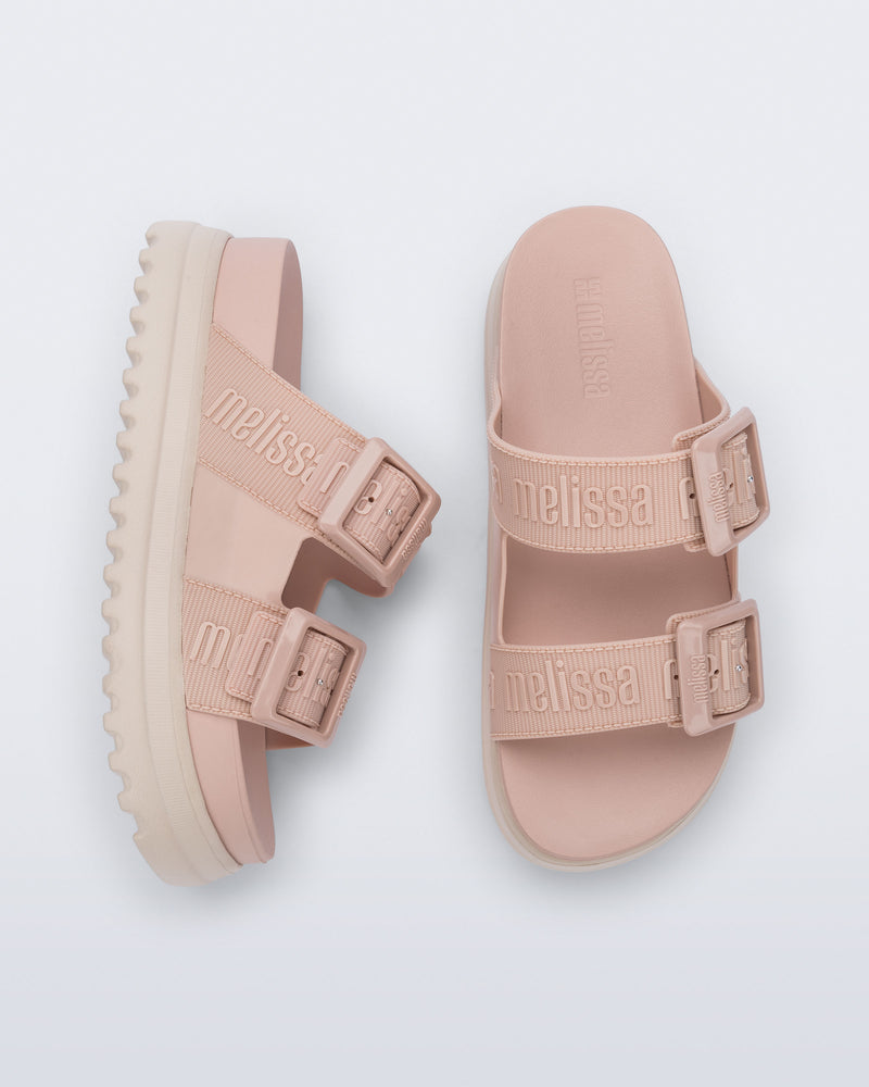 Top and side view of a pair of melissa cozy platform slide sandals. There are two buckle straps that go across the foot with the M Lover (melissa logo) print embossed into the straps. The buckles are located on the outside edge of the two straps. The straps and insole of the shoe are light pink and the platform sole is beige.