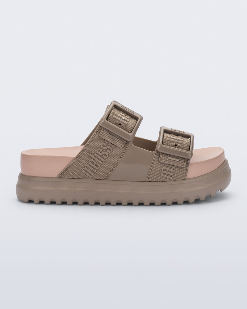 Side view of the melissa cozy platform slide sandal. There are two buckle straps that go across the foot with the M Lover (melissa logo) print embossed into the straps. The buckles are located on the outside edge of the two straps. The straps and platform sole of the shoe are dark beige color and the insole is a lighter color beige.