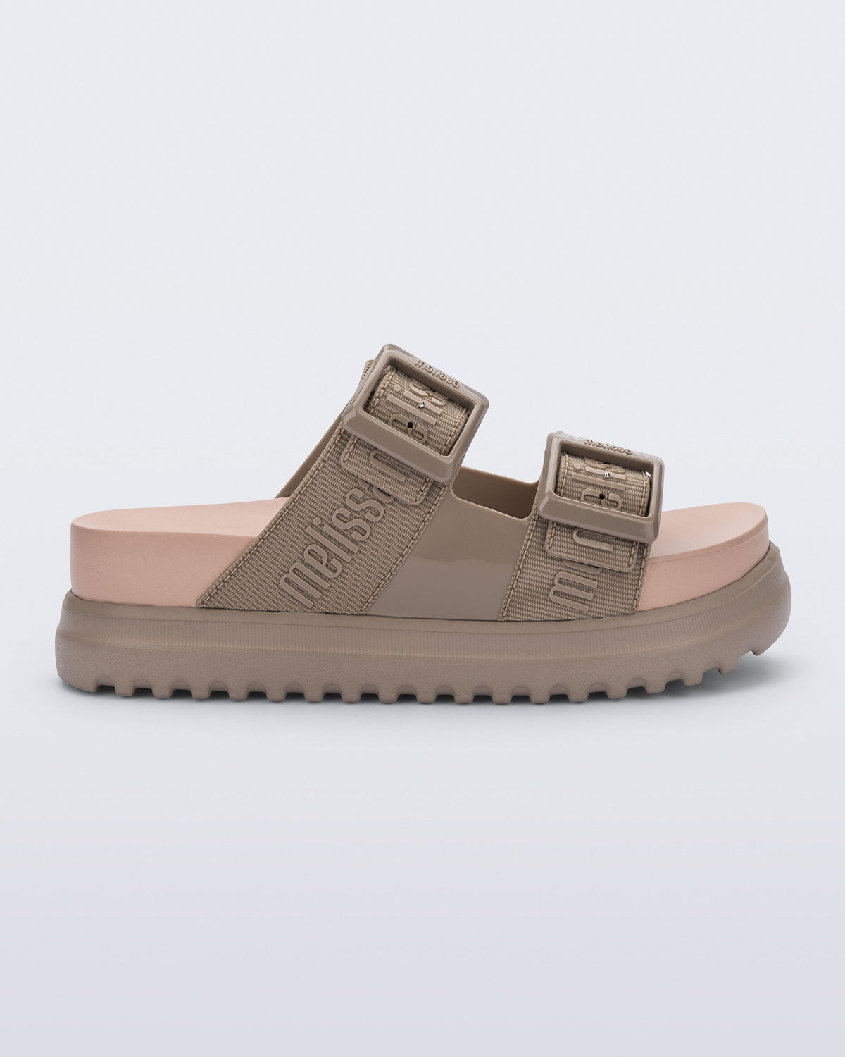 Side view of the melissa cozy platform slide sandal. There are two buckle straps that go across the foot with the M Lover (melissa logo) print embossed into the straps. The buckles are located on the outside edge of the two straps. The straps and platform sole of the shoe are dark beige color and the insole is a lighter color beige.