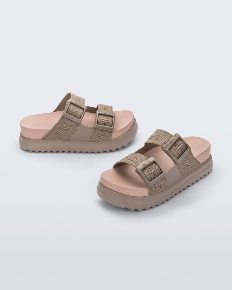 Angled view of a pair of melissa cozy platform slide sandals. There are two buckle straps that go across the foot with the M Lover (melissa logo) print embossed into the straps. The buckles are located on the outside edge of the two straps. The straps and platform sole of the shoe are dark beige color and the insole is a lighter color beige.