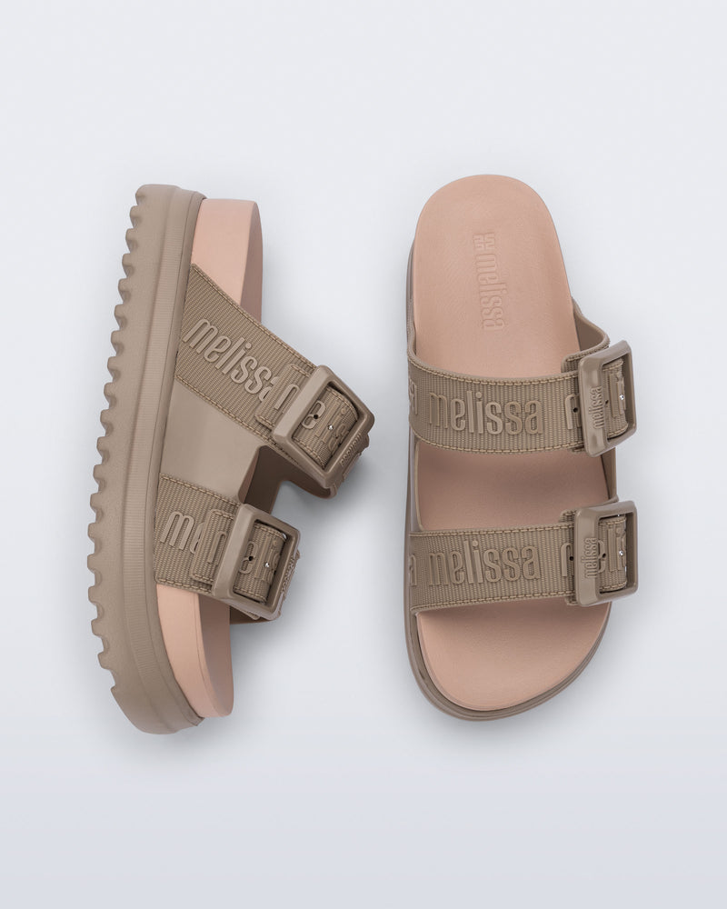 Top and side view of a pair of melissa cozy platform slide sandals. There are two buckle straps that go across the foot with the M Lover (melissa logo) print embossed into the straps. The buckles are located on the outside edge of the two straps. The straps and platform sole of the shoe are dark beige color and the insole is a lighter color beige.