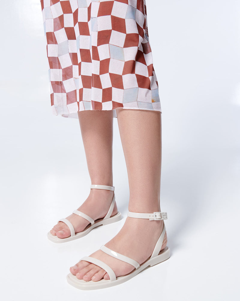 Close up view of a model's leggs posing in a pair of Melissa Heat Sandals. This is a flat sandal with a multiple straps, one across the toes, mid part of the foot, and a buckle strap around the ankle with a supporting strap that connects the ankle strap to the sole of the shoe. The straps, insole, outsole, and buckles on the ankle strap are beige. The insole is embossed with a geometric pattern.