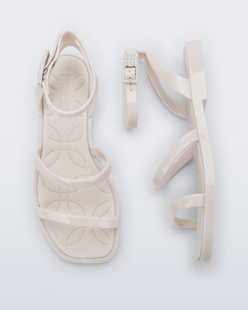 Top and side view of a pair of Melissa Heat Sandals. This is a flat sandal with a multiple straps, one across the toes, mid part of the foot, and a buckle strap around the ankle with a supporting strap that connects the ankle strap to the sole of the shoe. The straps, insole, outsole, and buckles on the ankle strap are beige. The insole is embossed with a geometric pattern.