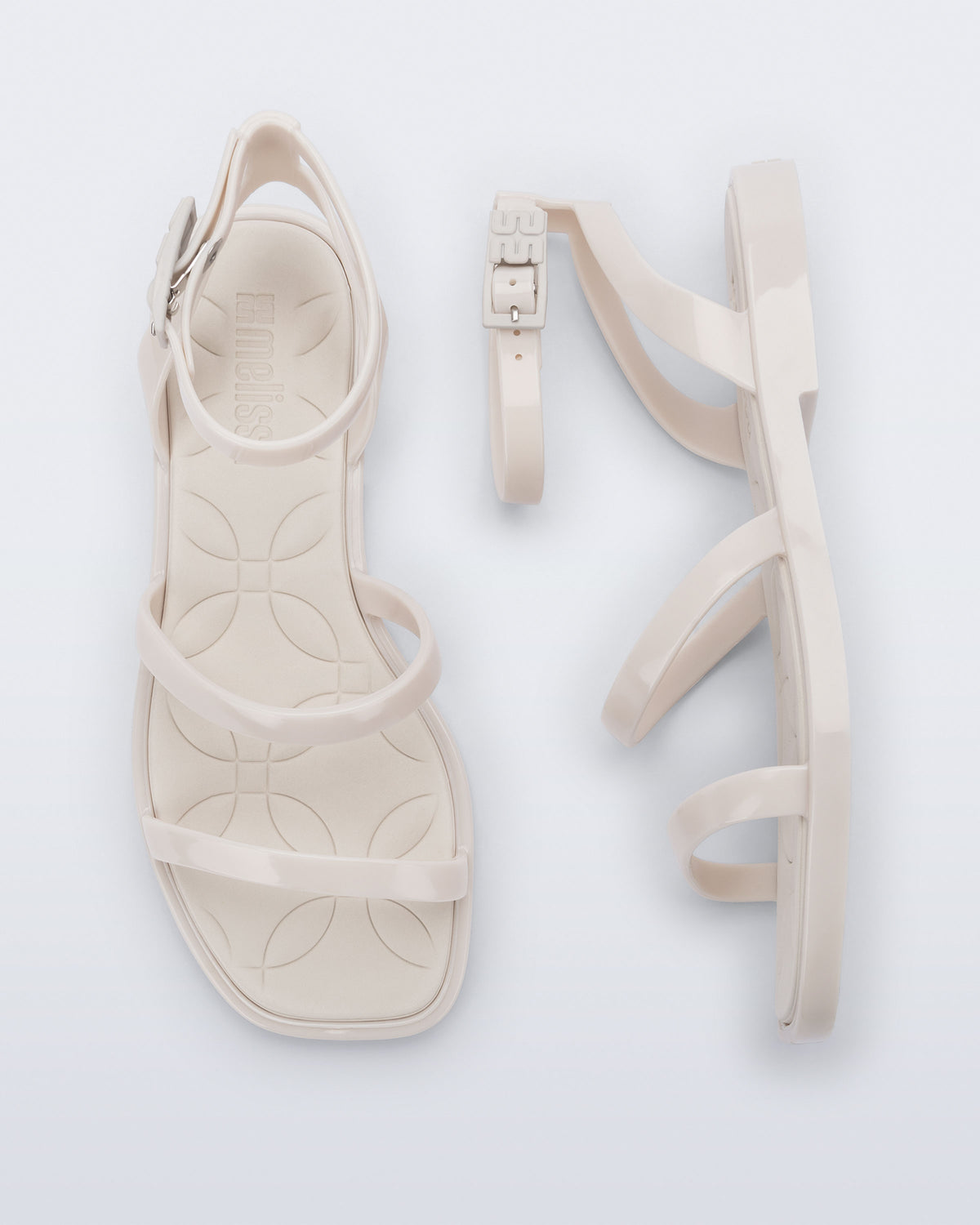 Top and side view of a pair of Melissa Heat Sandals. This is a flat sandal with a multiple straps, one across the toes, mid part of the foot, and a buckle strap around the ankle with a supporting strap that connects the ankle strap to the sole of the shoe. The straps, insole, outsole, and buckles on the ankle strap are beige. The insole is embossed with a geometric pattern.