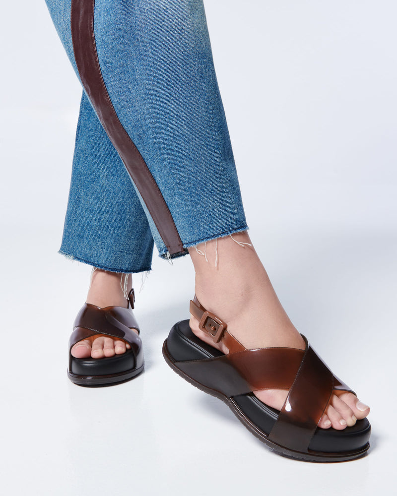 Close up of a model's legs posing in a pair of Melissa Mare platform sandals. This sandal has two cross straps across the front part of the foot with a thin buckle strap attached. The straps and outsole are brown while the platform insole is a black.