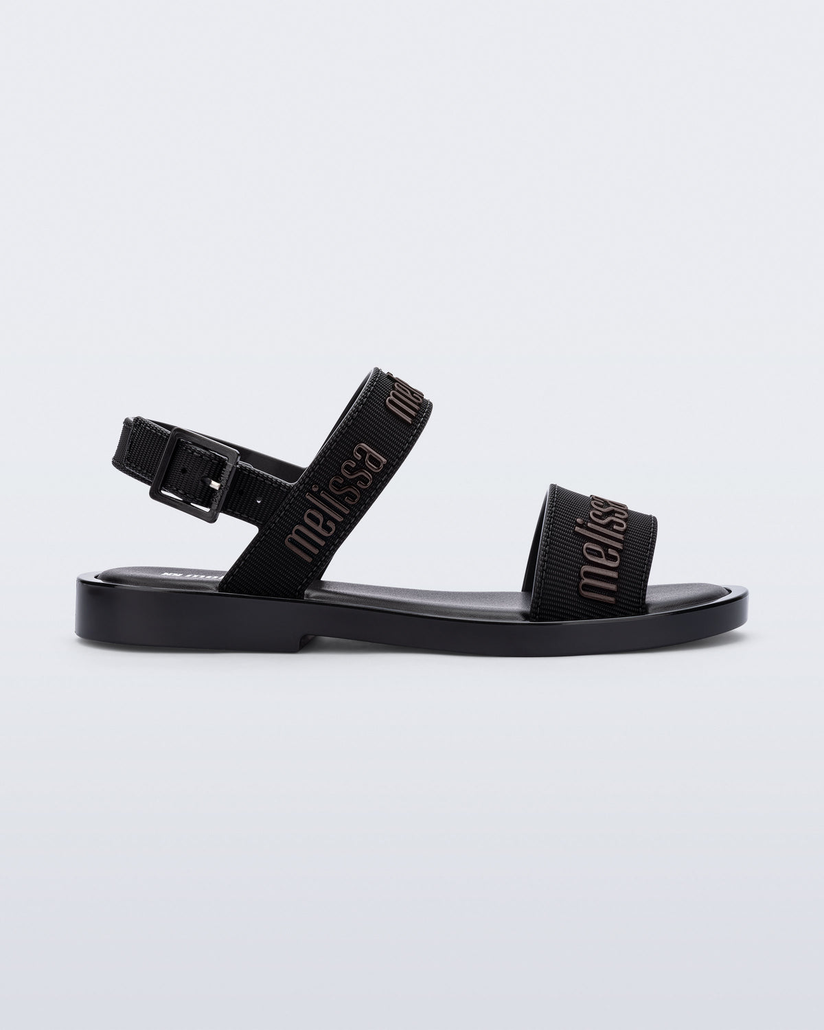 Side view of the Melissa Mar Sandal with Melissa logo embossed into the straps. There are two embossed straps that go across the foot; 1 across the toes and the other across the main part of the foot. The main foot strap has an ankle strap attached with a buckle closure. The straps ,buckle and sole of the shoe is black. The Melissa logo on the straps has a black metallic finish. . The buckle placement is on the outside of the ankle strap.