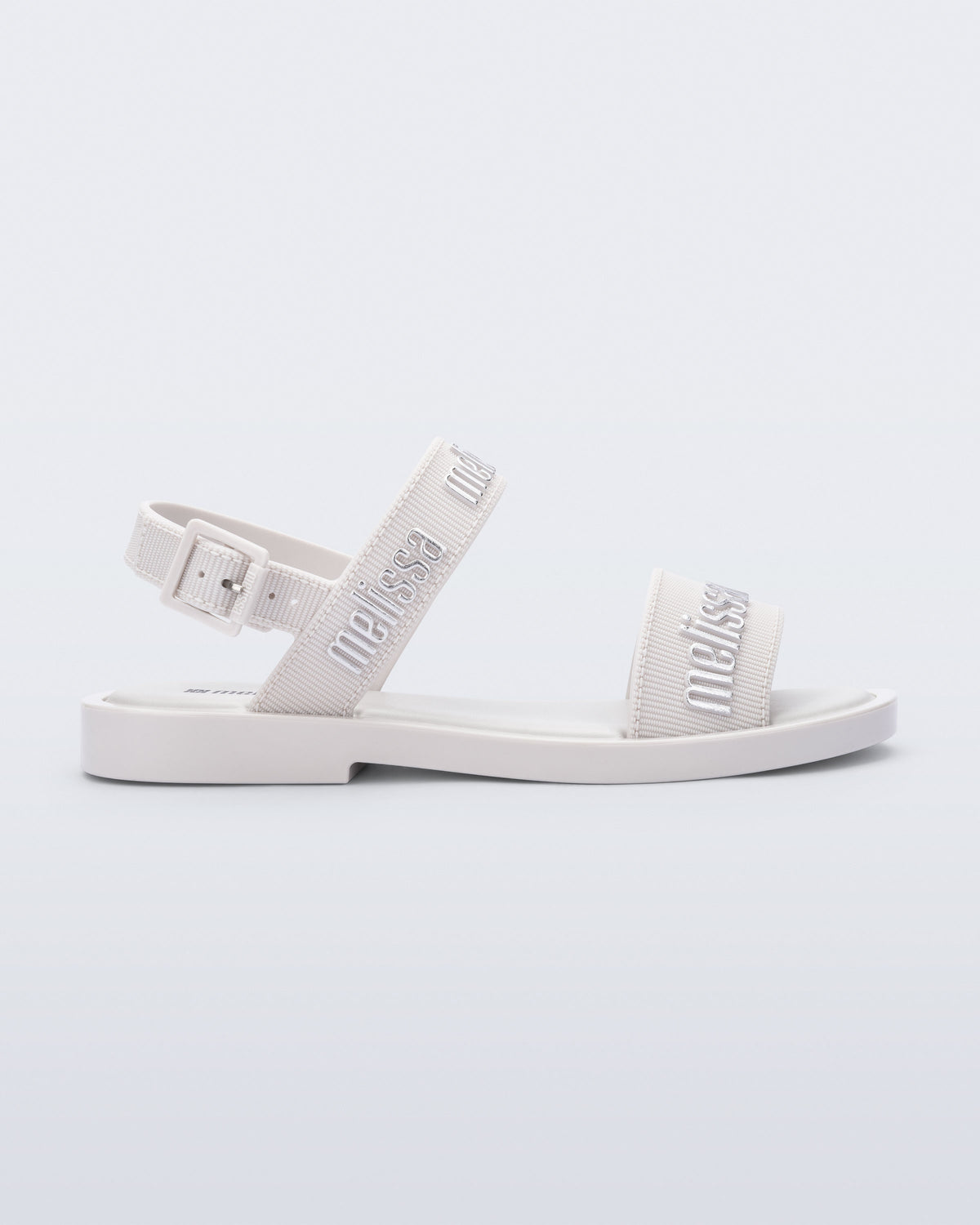 Side view of the Melissa Mar Sandal with Melissa logo embossed into the straps. There are two embossed straps that go across the foot; 1 across the toes and the other across the main part of the foot. The main foot strap has an ankle strap attached with a buckle closure. The straps ,buckle and sole of the shoe is white. The Melissa logo on the straps has a silver metallic finish. . The buckle placement is on the outside of the ankle strap.