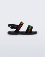 Side view of the Mini Melissa Mar Sandal with Mini Melissa logo embossed into the straps. There are two embossed straps that go across the foot; 1 across the toes and the other across the main part of the foot. The main foot strap has an ankle strap attached with a buckle closure. The straps ,buckle and sole of the shoe is black. The Melissa logo on the straps has a rainbow metallic finish. . The buckle placement is on the outside of the ankle strap.