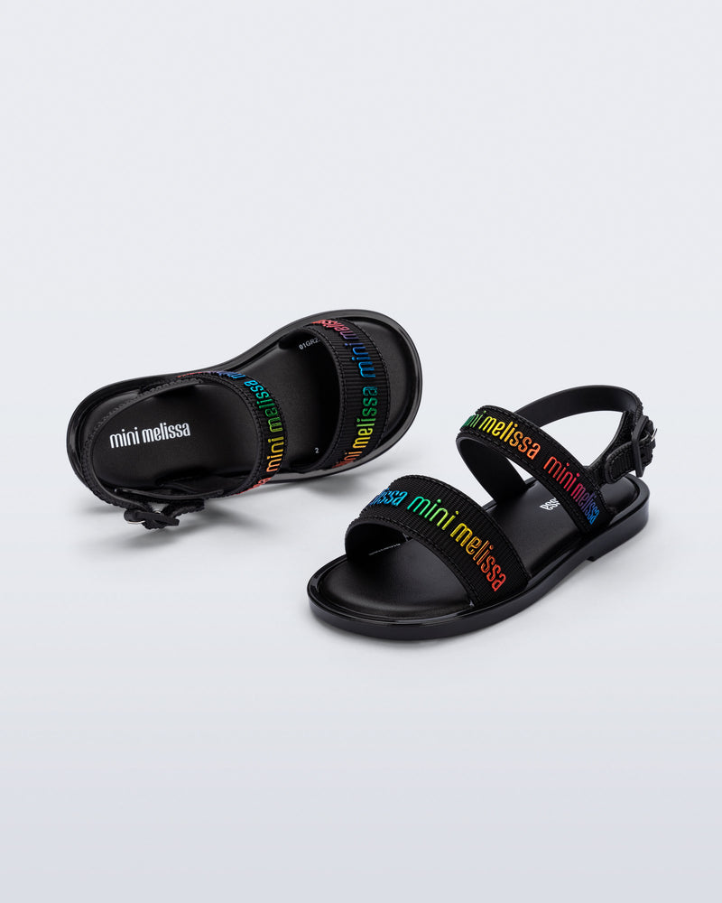 Angled view of a pair of Mini Melissa Mar Sandals with Mini Melissa logo embossed into the straps. There are two embossed straps that go across the foot; 1 across the toes and the other across the main part of the foot. The main foot strap has an ankle strap attached with a buckle closure. The straps ,buckle and sole of the shoe is black. The Melissa logo on the straps has a rainbow metallic finish. The buckle placement is on the outside of the ankle strap.