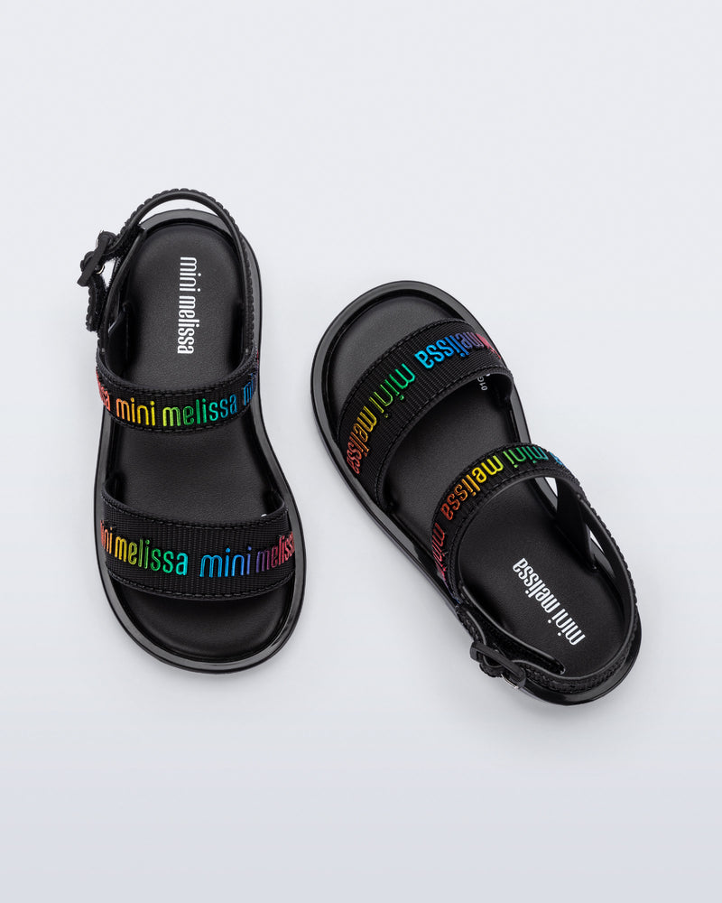 Top view of a pair of Mini Melissa Mar Sandals with Mini Melissa logo embossed into the straps. There are two embossed straps that go across the foot; 1 across the toes and the other across the main part of the foot. The main foot strap has an ankle strap attached with a buckle closure. The straps ,buckle and sole of the shoe is black. The Melissa logo on the straps has a rainbow metallic finish. The buckle placement is on the outside of the ankle strap.