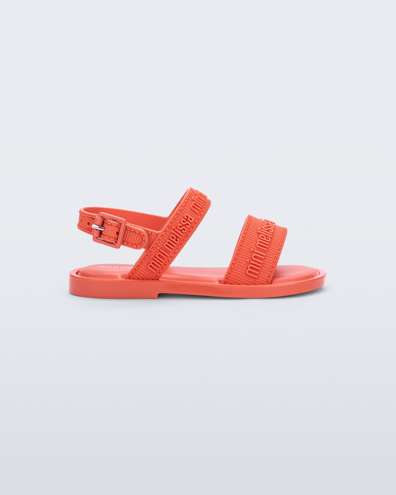 Side view of the Mini Melissa Mar Sandal with Mini Melissa logo embossed into the straps. There are two embossed straps that go across the foot; 1 across the toes and the other across the main part of the foot. The main foot strap has an ankle strap attached with a buckle closure. The buckle placement is on the outside of the ankle strap. The color of the shoe is orange.