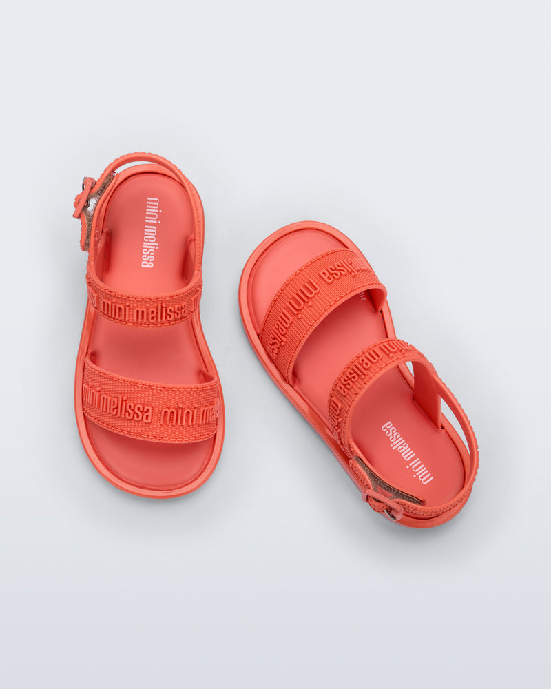 Top view of a pair of Mini Melissa Mar Sandals with Mini Melissa logo embossed into the straps. There are two embossed straps that go across the foot; 1 across the toes and the other across the main part of the foot. The main foot strap has an ankle strap attached with a buckle closure. The color of the shoe is orange. The buckle placement is on the outside of the ankle strap.