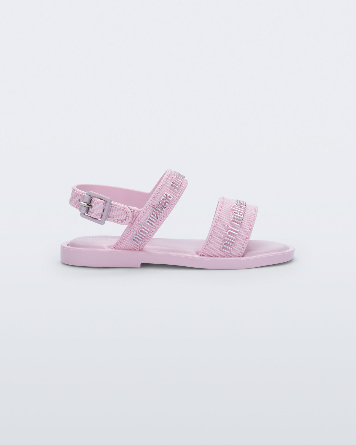 Side view of the Mini Melissa Mar Sandal with Mini Melissa logo embossed into the straps. There are two embossed straps that go across the foot; 1 across the toes and the other across the main part of the foot. The main foot strap has an ankle strap attached with a buckle closure. The straps ,buckle and sole of the shoe is pink. The Melissa logo on the straps has a silver metallic finish. . The buckle placement is on the outside of the ankle strap.