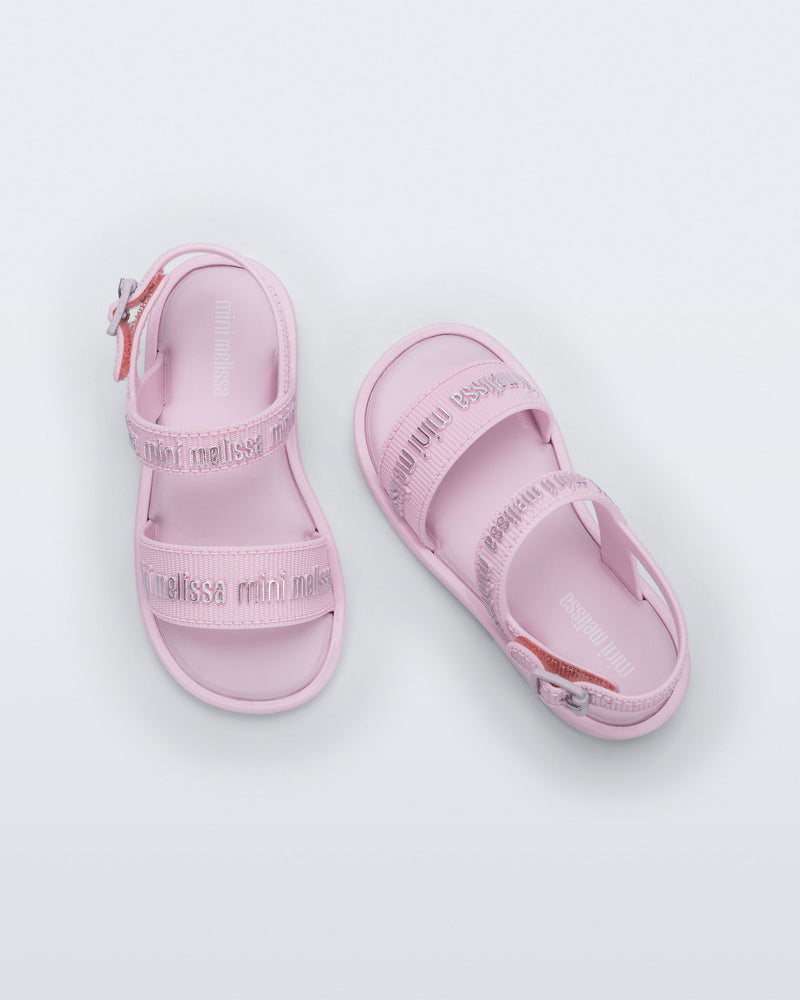 Top view of a pair of Mini Melissa Mar Sandals with Mini Melissa logo embossed into the straps. There are two embossed straps that go across the foot; 1 across the toes and the other across the main part of the foot. The main foot strap has an ankle strap attached with a buckle closure. The straps ,buckle and sole of the shoe is pink. The Melissa logo on the straps has a silver metallic finish. The buckle placement is on the outside of the ankle strap.