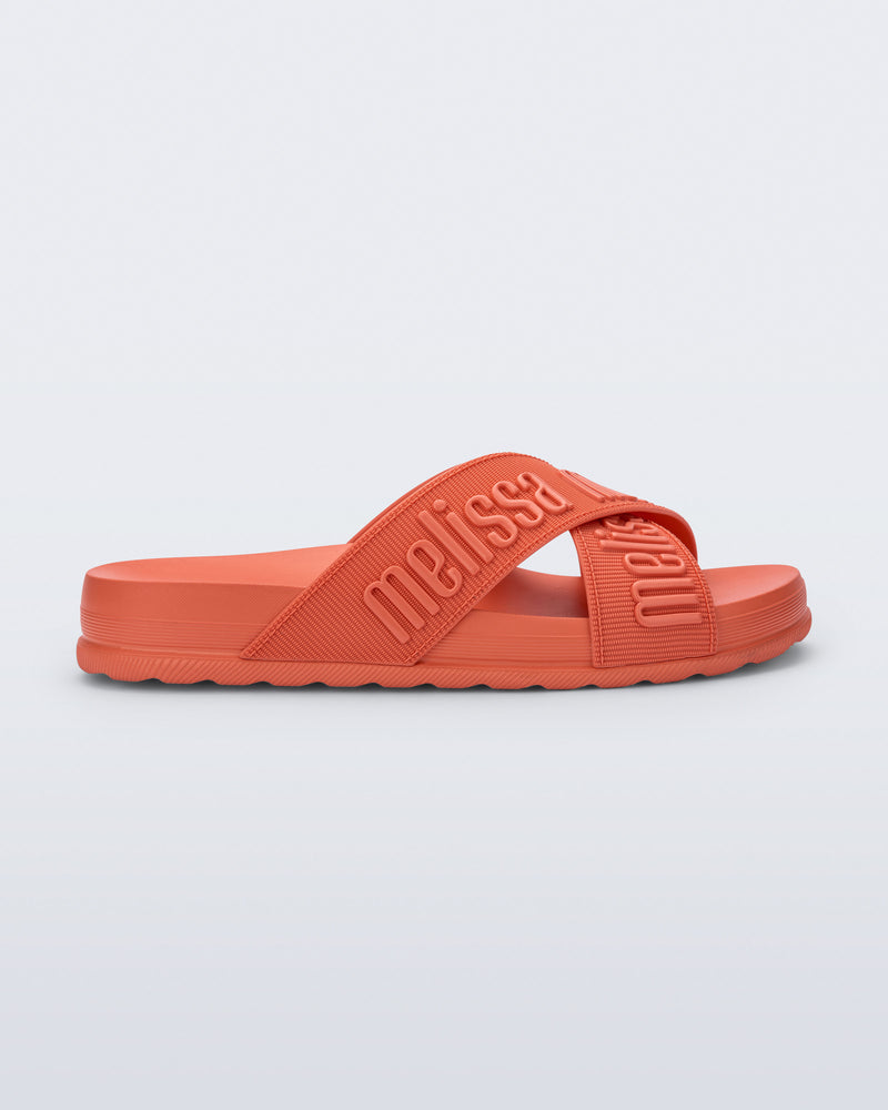 Side view of the Melissa Cozy slide sandals. The slide has two cross straps with Melissa logo embossed onto the straps. The color of the shoe is orange.