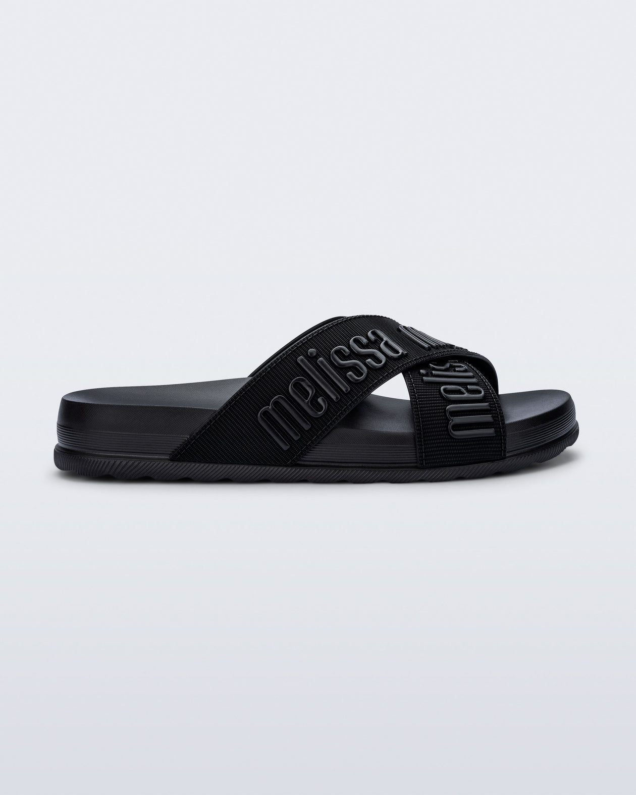 Side view of the Melissa Cozy slide sandals. The slide has two cross straps with Melissa logo embossed onto the straps. The color of the shoe is black.