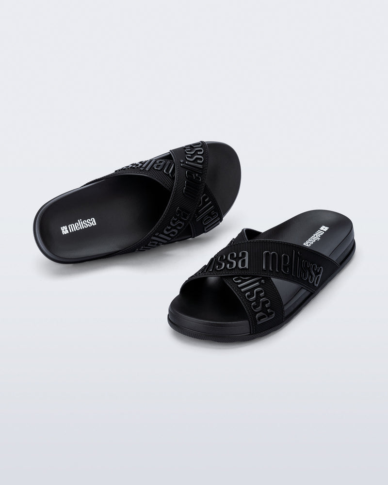 Angled view of a pair of Melissa Cozy slide sandals. The slide has two cross straps with Melissa logo embossed onto the straps. The color of the shoe is black.