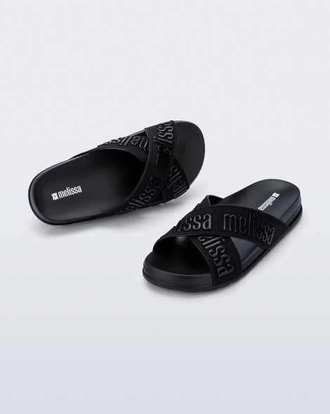 Angled view of a pair of Melissa Cozy slide sandals. The slide has two cross straps with Melissa logo embossed onto the straps. The color of the shoe is black.