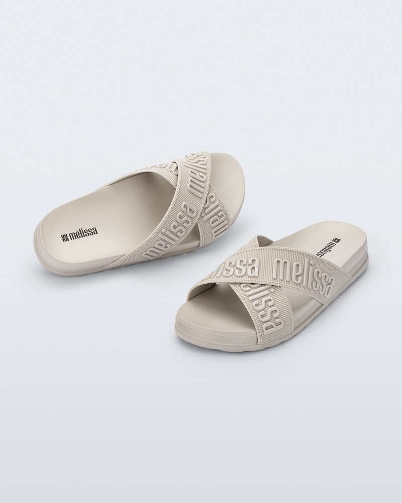 Angled view of a pair of Melissa Cozy slide sandals. The slide has two cross straps with Melissa logo embossed onto the straps. The color of the shoe is beige.