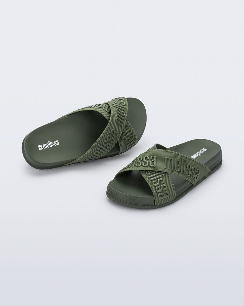 Angled view of a pair of Melissa Cozy slide sandals. The slide has two cross straps with Melissa logo embossed onto the straps. The color of the shoe is a dark green.