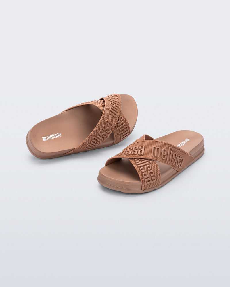 Angled view of a pair of Melissa Cozy slide sandals. The slide has two cross straps with Melissa logo embossed onto the straps. The color of the shoe is brown.