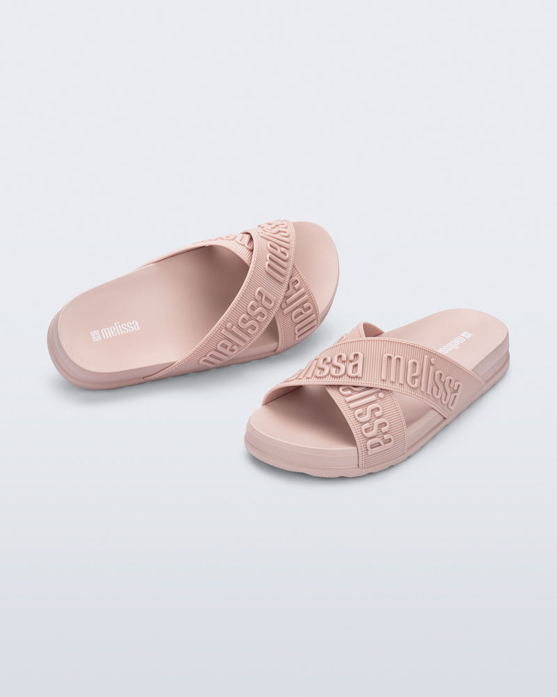 Angled view of a pair of Melissa Cozy slide sandals. The slide has two cross straps with Melissa logo embossed onto the straps. The color of the shoe is light pink.