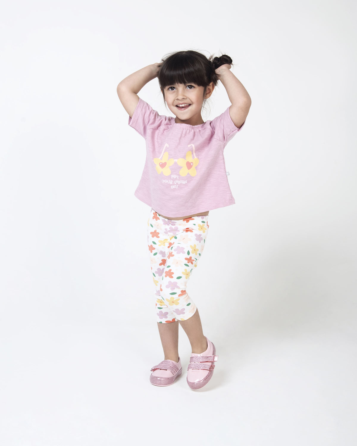 A model posing for a picture in a pink graphic tee, white patterend pants and a pair of Pink/Pink Glitter Mini Melissa Beanny Bugs sneakers with a pink base, two shiny pink velcro straps and a pink glitter sole.