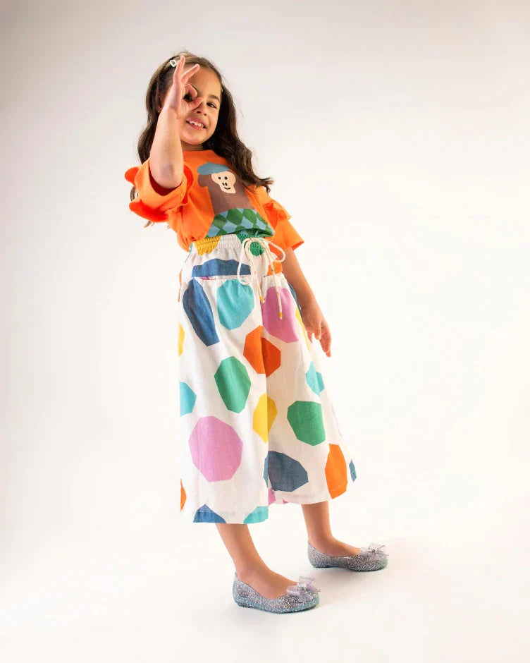 A young model posing for a picture in an orange top, white patterned pants, and a pair of clear Mini Melissa Sweet Love Princess flats, with a clear glitter base, Elsa in script on the side, a bow detail on the toe, and a drawing of Elsa of Frozen on the sole.