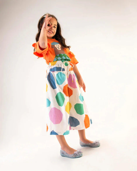 A young model posing for a picture in an orange top, white patterned pants, and a pair of clear Mini Melissa Sweet Love Princess flats, with a clear glitter base, Elsa in script on the side, a bow detail on the toe, and a drawing of Elsa of Frozen on the sole.
