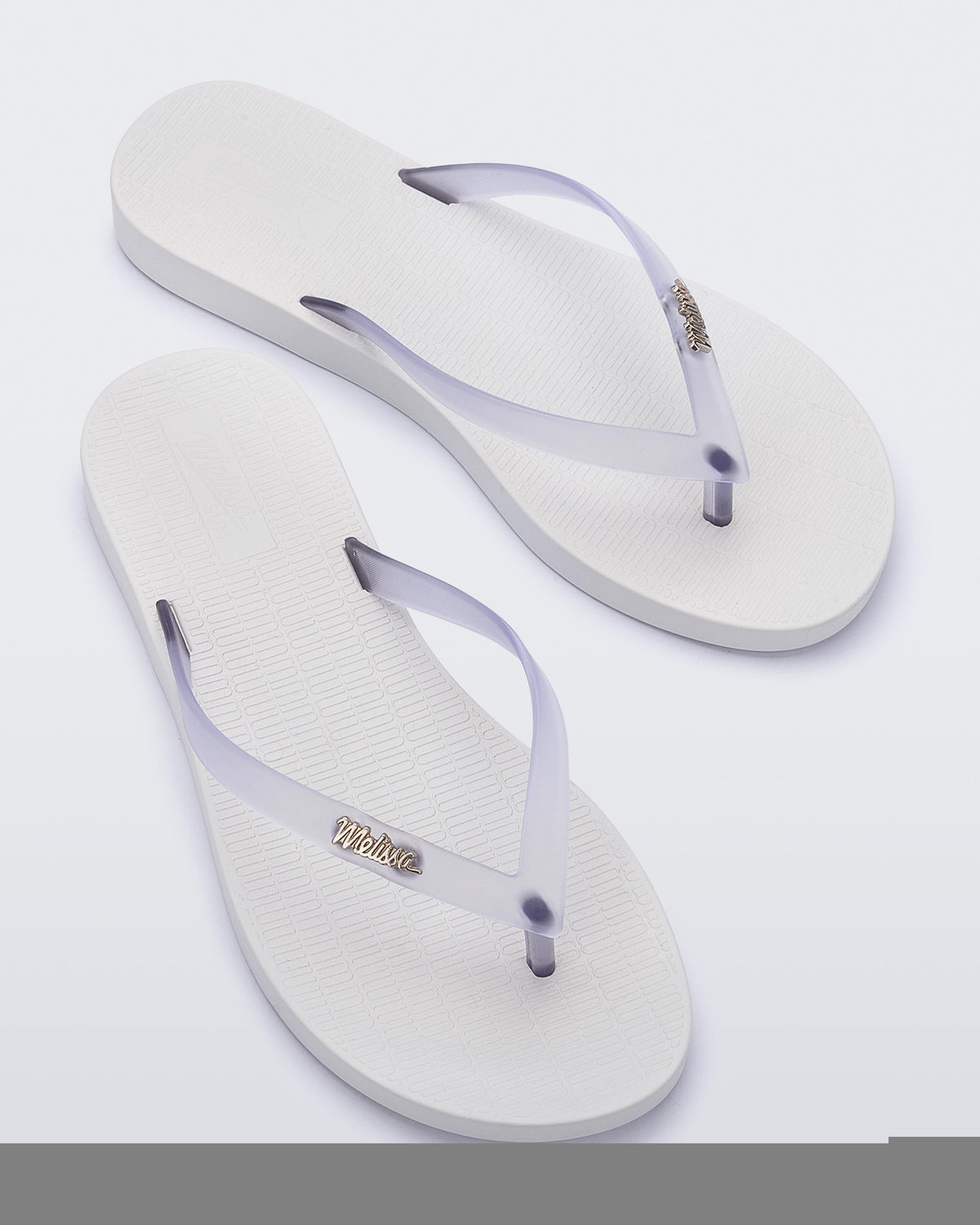 Top view of a pair of white/clear Melissa Sun Venice flip flops with a white sole and clear straps with "melissa" on one strap.