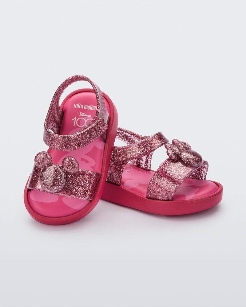 An angled top and side view of a pair of glitter pink Mini Melissa Jump sandals, leaning on eachother, with a Mickey Mouse logo detail on the front strap and an ankle strap