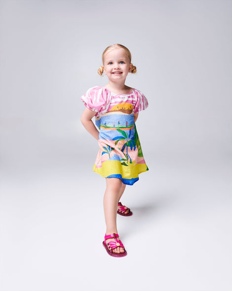 Child model in a multi colored dress wearing a pair of glitter pink baby Hip Bloomy sandal with pink straps and flower